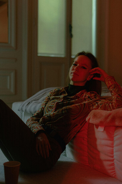 A woman sits on the floor, leaning against her bed in a dark room, lost in quiet reflection