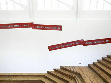 The artwork shown in the image is a conceptual wall installation by Lawrence Weiner titled laid out on the banks of a river / leaning towards the ocean / leaning towards the land (2017), commissioned specifically for the Art Wall at the Berkeley Art Museum and Pacific Film Archive