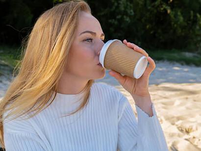 A happy woman sips coffee from a paper cup, wearing headphones for audio healing, uniting wellbeing, nature, and mindfulness.