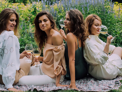 Underneath the open sky, four women sit on the lush green grass, sharing laughter and clinking wine glasses as they celebrate the camaraderie of Galentine's Day, cherishing the bonds of their enduring friendship