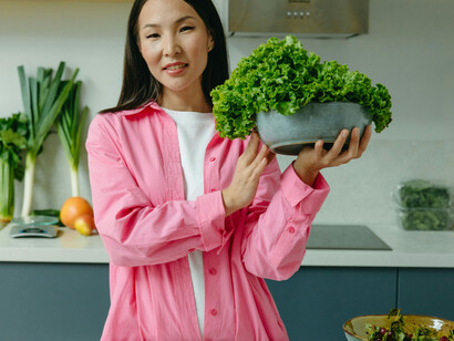 A woman in the kitchen filled with produce wearing a pink shirt holding a bowl of vegetables