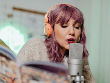 A woman wearing headphones records a podcast indoors while reading from a book in a relaxed and creative setting