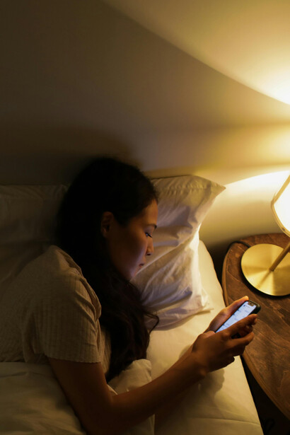A woman lies on her bed in a dimly lit room, illuminated by the glow of her phone screen