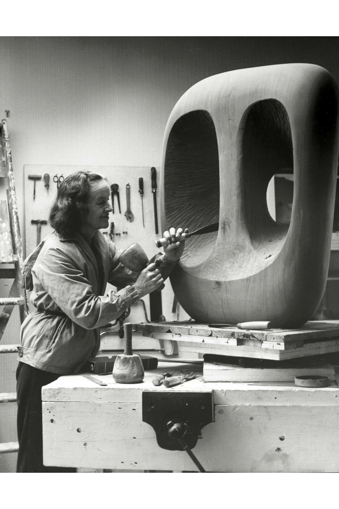 Barbara Hepworth in the Palais studio in with unfinished wood carving Hollow Form with White Interior, 1963, Photograph: Val Wilmer, Courtesy © Bowness, Hepworth Estate
