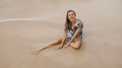 A cheerful and radiant woman sits on the sandy beach, wearing a smile that brightens the surroundings