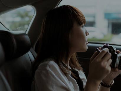 A serene woman, reclining in the car's backseat, gazes out of the window as they move away, her wistful expression complemented by the presence of a photo camera nearby