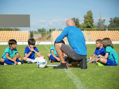 A sports coach in a blue shirt mentoring a group of children while giving instructions on a soccer field