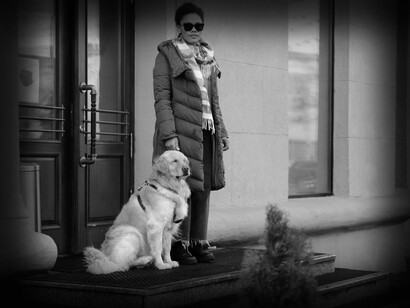 A blind woman stands on her doorstep with her seeing-eye-dog