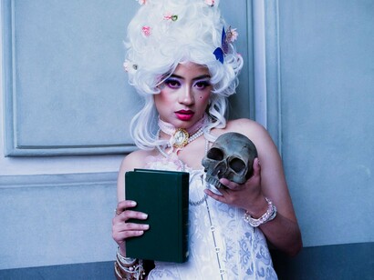 Uma mulher em vestido de inspiração vintage posa com uma caveira e um livro. A composição aproxima teatro, literatura e passagem do tempo, evocando o universo de Shakespeare e a permanência de suas obras através dos séculos