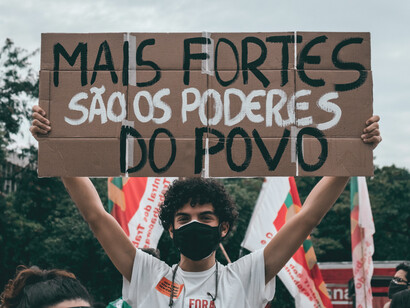 Un joven manifestante alzando con ambas manos un cartel que dice "Más fuertes son los poderes del pueblo" frente a banderas de movimientos sociales