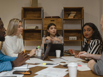 In a boardroom setting, a woman in a gray blazer delivers a report and takes part in a collaborative business discussion