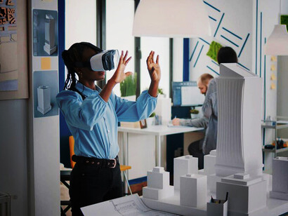 Architect wearing VR goggles views a 3D plan of a white foam maquette in a modern office, while an engineer works with a VR headset next to a table with an urban project model