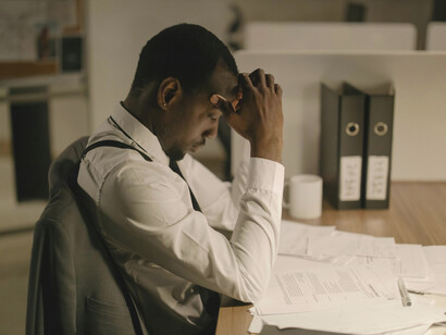 A man works late in an office surrounded by stacks of paperwork and files