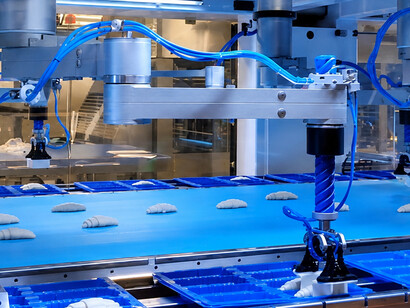 Mechanical robots replace manual labor by moving semi-finished French croissants from conveyor belts into containers during large-scale production