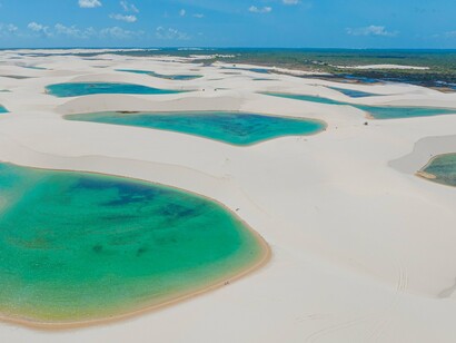 Uma lagoa de águas verdes e azuladas ocupa o primeiro plano, cercada por extensas dunas de areia clara. A imagem remete ao encontro entre água e areia que encanta o narrador e transforma a caminhada em uma experiência de contemplação