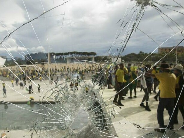 Por trás do vidro estilhaçado, manifestantes vestidos predominantemente de verde e amarelo ocupam a rampa de acesso ao Palácio do Planalto, durante os atos de 8 de janeiro de 2023. A destruição das sedes dos Três Poderes expôs a gravidade da ofensiva contra as instituições democráticas brasileiras perpetradas por estes mesmos representantes da extrema-direita