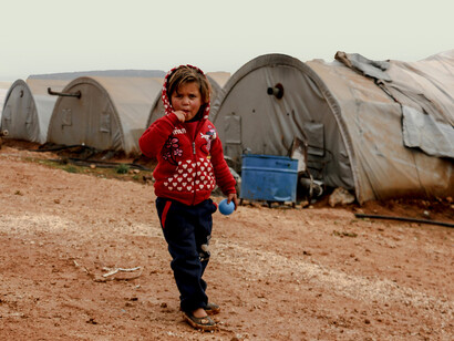 A young girl walks through a desert refugee camp, surrounded by tents under the harsh sun