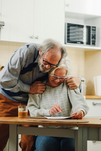 An elderly couple reading a document in their kitchen, laughing together in a moment that reflects the strength of a long-term relationship