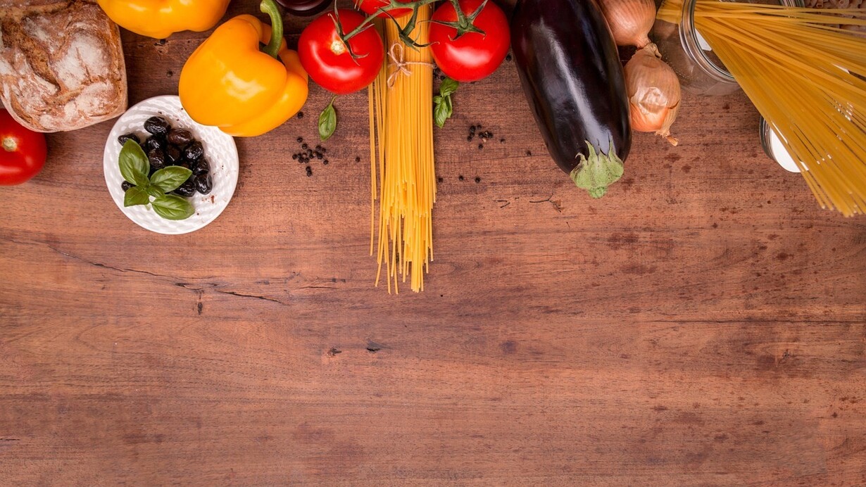 A table showing an assorted selection of food, including pasta, tomatoes, eggplants, bell peppers, onions, basil, olives and bread
