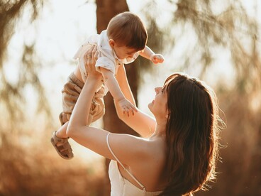 A mother joyfully lifting her baby into the sunlight, representing the secure attachment that lays the foundation for lifelong emotional well-being