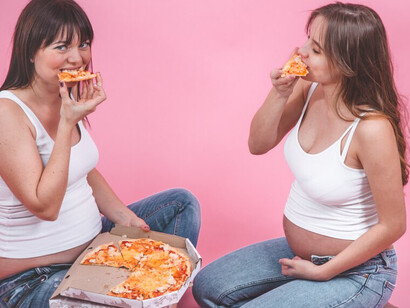 regnant women eating pizza against a pink wall, illustrating Western diet foods and the health risks associated with processed foods