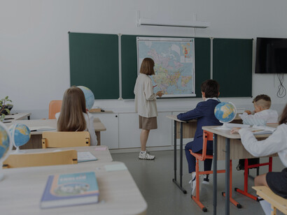 Una docente de primaria con ropas de colores neutros dando una clase en el pizarrón, de espaldas a sus estudiantes