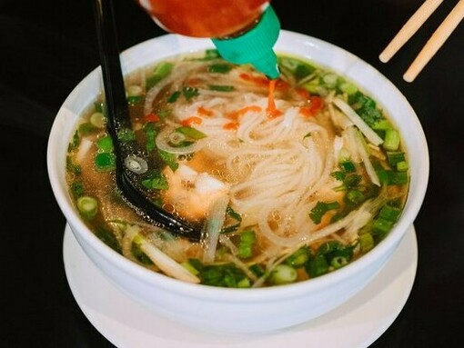 A steaming bowl of pho, a classic Vietnamese noodle soup with delicate rice noodles and fresh herbs, is being enhanced with a drizzle of sriracha, adding a touch of spice to this comforting and flavorful dish
