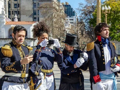 Personagens vestidos com trajes históricos participam de uma reconstituição em Buenos Aires, enquanto a tecnologia contemporânea registra a cena. O contraste remete à distância que pode existir entre a imagem construída durante uma campanha e a realidade vivida depois da eleição. Em setembro de 2026, a pobreza na Argentina chegou a 32,3% da população no primeiro semestre, segundo dados oficiais, em meio às dificuldades sociais associadas ao ajuste econômico do governo Milei