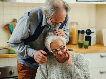 An elderly couple sits in the kitchen, reading a document and laughing together, illustrating the comfort and connection of a long-term relationship