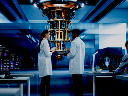 A young female team leader and a male Indian scientist stand in a futuristic research center, discussing a quantum computing device while observing the laboratory from behind