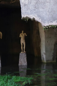 Abandoned and neglected sculptures stand in a cave beside a still body of water, remnants of a forgotten artistic legacy