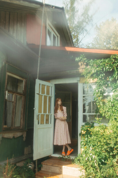 A young woman in a vintage dress pauses at the entrance of a rustic country house, enveloped by rich greenery, blooming flowers, and gentle summer light