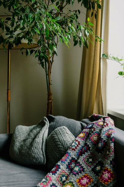Um sofá, uma manta colorida e plantas ocupam uma sala iluminada, compondo um ambiente de aspecto acolhedor e vivido. A casa aparece aqui menos como cenário e mais como espaço de conforto, memória e experiências cotidianas