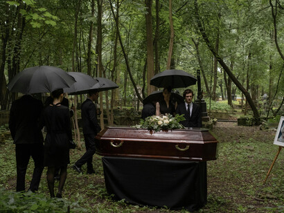 In the forest, people in black gather around a brown coffin for a funeral service