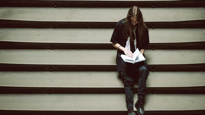 A person sitting on a set of stairs, holding a book, lost in thought or overwhelmed by the weight of their studies