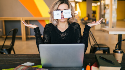 A portrait of a young woman sitting at a table, paper stickers stuck to her glasses, her expression tired and worn