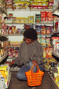 An African American woman selects products while shopping in a convenience store, carrying an orange shopping basket