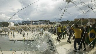Por trás do vidro estilhaçado, manifestantes vestidos predominantemente de verde e amarelo ocupam a rampa de acesso ao Palácio do Planalto, durante os atos de 8 de janeiro de 2023. A destruição das sedes dos Três Poderes expôs a gravidade da ofensiva contra as instituições democráticas brasileiras perpetradas por estes mesmos representantes da extrema-direita