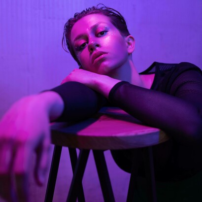 Primo piano di una persona appoggiata su uno sgabello di legno, illuminata da una luce viola e rosa neon