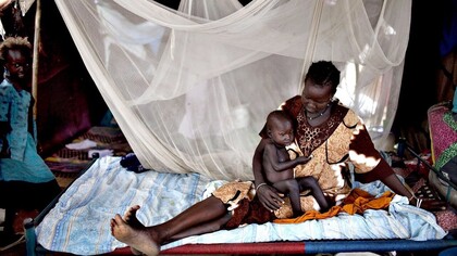 A Nilote woman laying with her baby son and her little daughter standing close by the bed, Africa