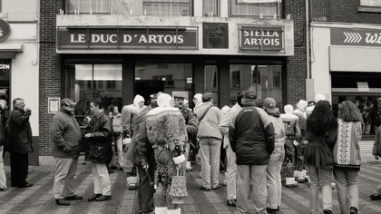 A black-and-white photograph of people standing in a queue outside a shop, Belgium