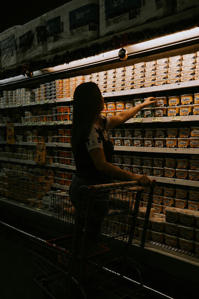 A silhouetted woman pushes a shopping cart through a dark grocery aisle lined with illuminated shelves of product