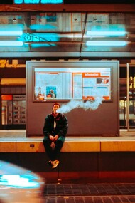 A man sitting at a bus stop and vaping, reflecting how people often turn to vaping as a way to cope with stress or anxiety in an increasingly hectic world