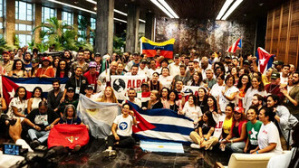 Integrantes da ALBA Movimentos reúnem-se durante a 4ª Assembleia Continental da organização, em Havana, diante das bandeiras de Cuba, Venezuela e Argentina. A imagem traduz a articulação política entre movimentos populares latino-americanos que o artigo apresenta como parte da disputa pela soberania e contra a ingerência imperial na região. A diversidade de movimentos representada na cena traduz o projeto de integração popular e solidariedade internacionalista que está no centro da articulação. A Alba Movimentos (Articulação Continental de Movimentos Sociais e Populares rumo à Alba) é uma rede que une centenas de organizações populares da América Latina e do Caribe — como sindicatos, grupos feministas, movimentos estudantis, camponeses e indígenas. Inspirada na aliança de governos da ALBA-TCP, ela atua de forma autônoma na base da sociedade para promover a integração regional e a resistência anticapitalista