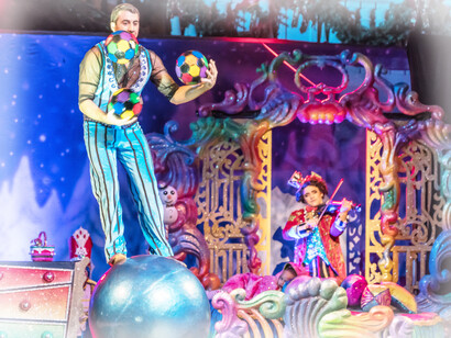 A juggler elevates his act, standing on a ball to continue his juggling while the other performer plays the violin along side the event