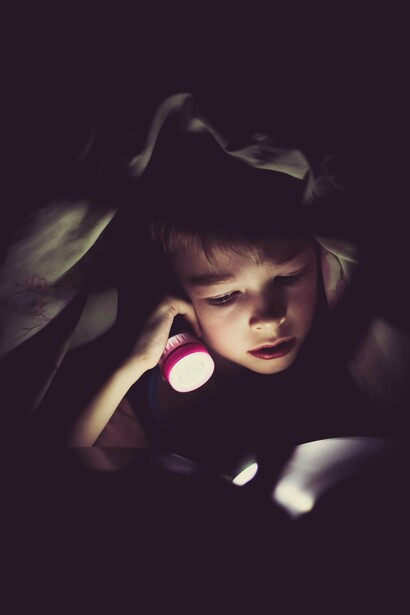 A child lying in bed, using a flashlight to read a comic book