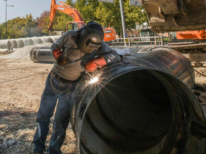 A man wearing a black jacket and blue denim jeans sits on a pipeline, performing repairs during the daytime in Turkey