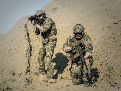 Dois soldados equipados para operações em campo aparecem em um ambiente árido, onde a movimentação e a adaptação ao terreno fazem parte das condições de atuação militar. Em guerras prolongadas, fatores como treinamento, logística e capacidade operacional influenciam a sustentação dos combates e as possibilidades de avanço ou recuo das forças envolvidas