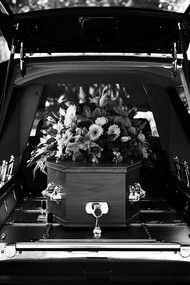 A grayscale photo of a car adorned with flowers, part of a solemn funeral procession