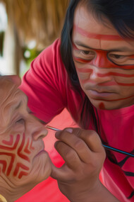 O artista Jaider Esbell pintando rosto de uma senhora de sua comunidade indígena em Roraima, Brasil